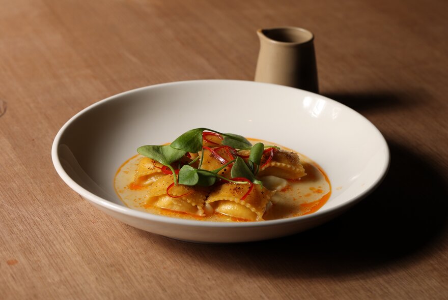 A bowl of ravioli with basil garnish and a small vessel behind it. 