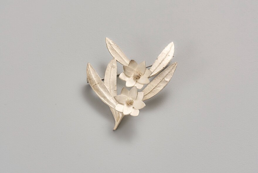 A delicate silver floral brooch featuring textured leaves and double floral detail, against a grey background. 