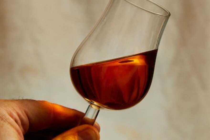 Hand holding a tulip-shaped glass of whisky, tilted to the side.
