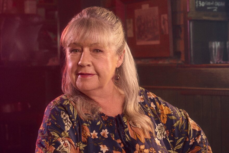 Actor Noni Hazelhurst, with a slight smile and her air pulled back from the sides of her face in a deep burgundy room