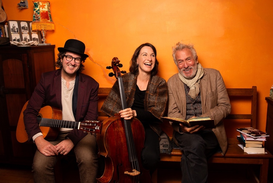 Two musicians and a man with a book in an orange room.
