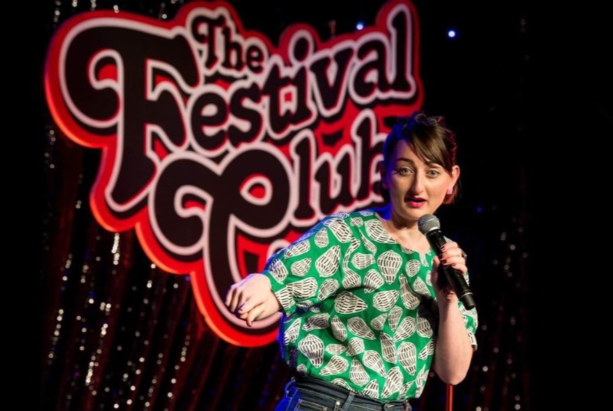 Comedian Sonia Di Iorio performing holding a microphone on stage in front of signage.