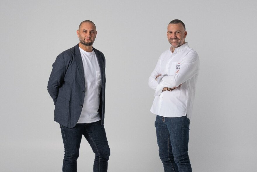 Two men smiling and looking at the camera, in front of a grey background.