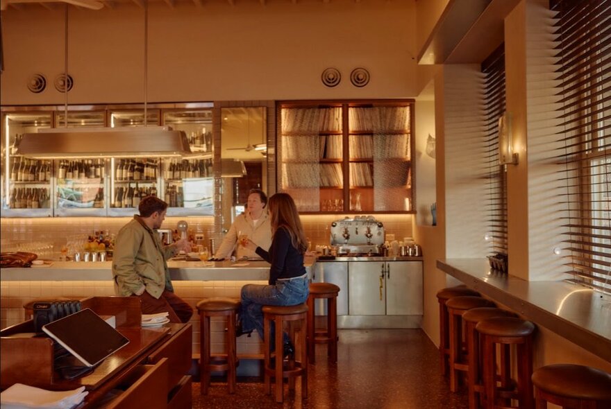 Warm bistro bar interior with couple seated on stools at a bench with host, sharing drinks and food, shelves of produce and wine in the background.