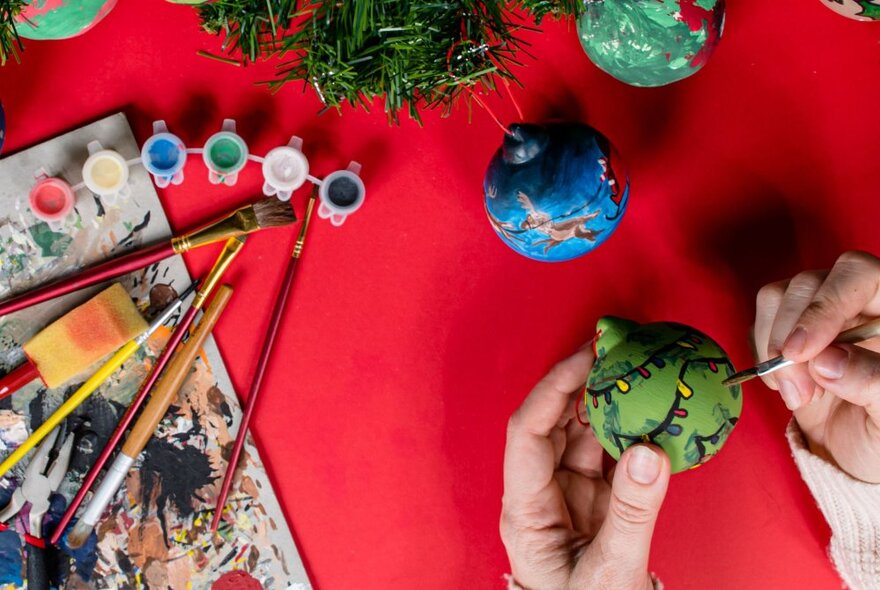 Looking down at a hand painting intricate decorations on a clay bauble, with other painted baubles, brushes and paints arranged on the table.