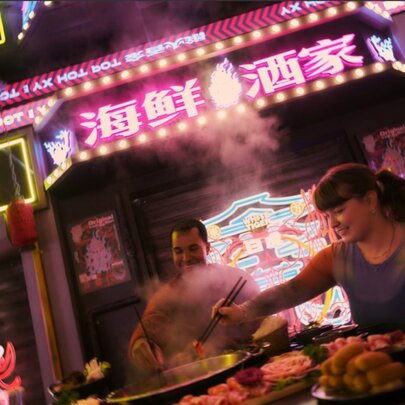 Diners using chopticks to choose food from an array of dishes in a neon-lit Chinese restaurant.