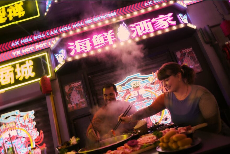 Diners using chopticks to choose food from an array of dishes in a neon-lit Chinese restaurant.