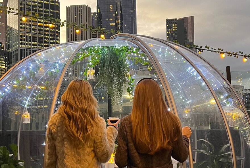 Two women seen from behind as they enter a rooftop glass dome with Melbourne's city buildings behind it.