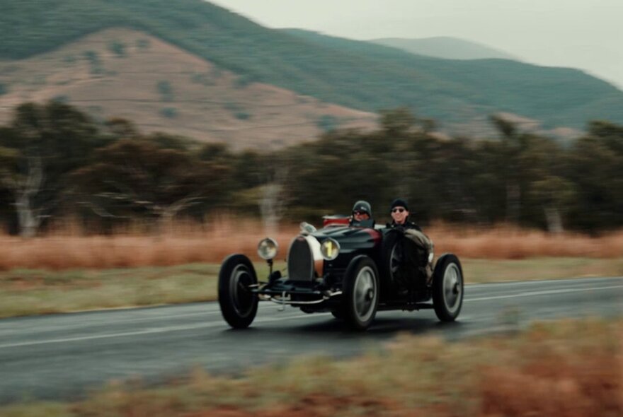 Vintage roadster being driven along a country road with hills and trees.