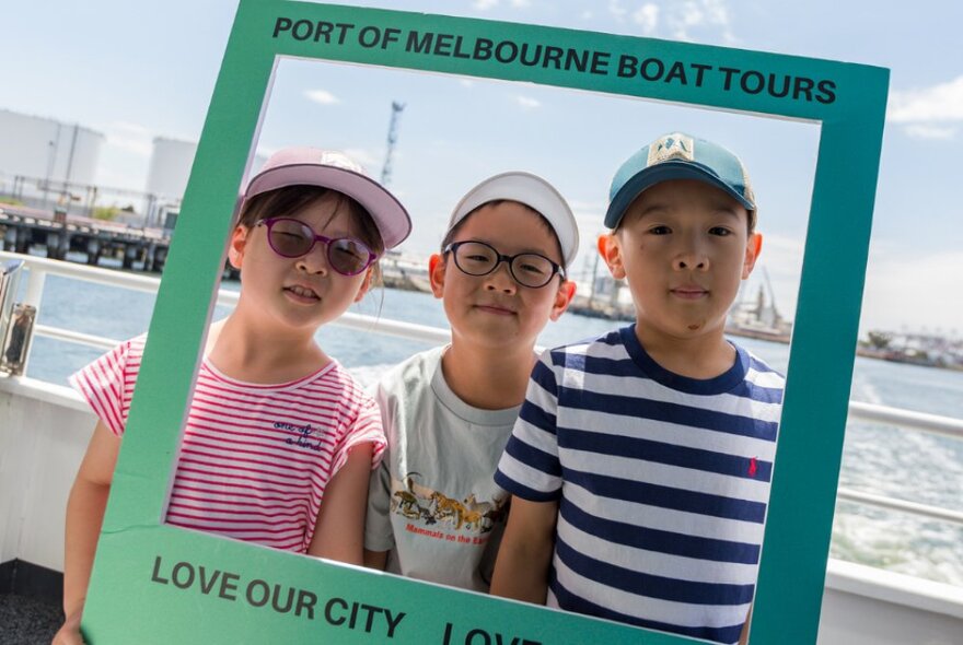 Three young children posing for a photo behind a temporary cardboard frame that reads PORT OF MELBOURNE BOAT TOURS, with a view of the port and river in the background.