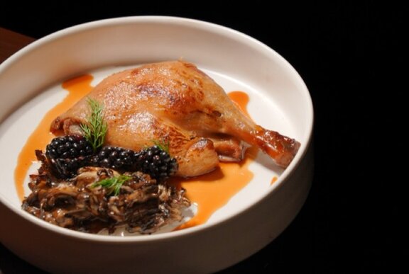A roast duck or chicken leg served with a side of dark grains and a rich sauce in a shallow white bowl.