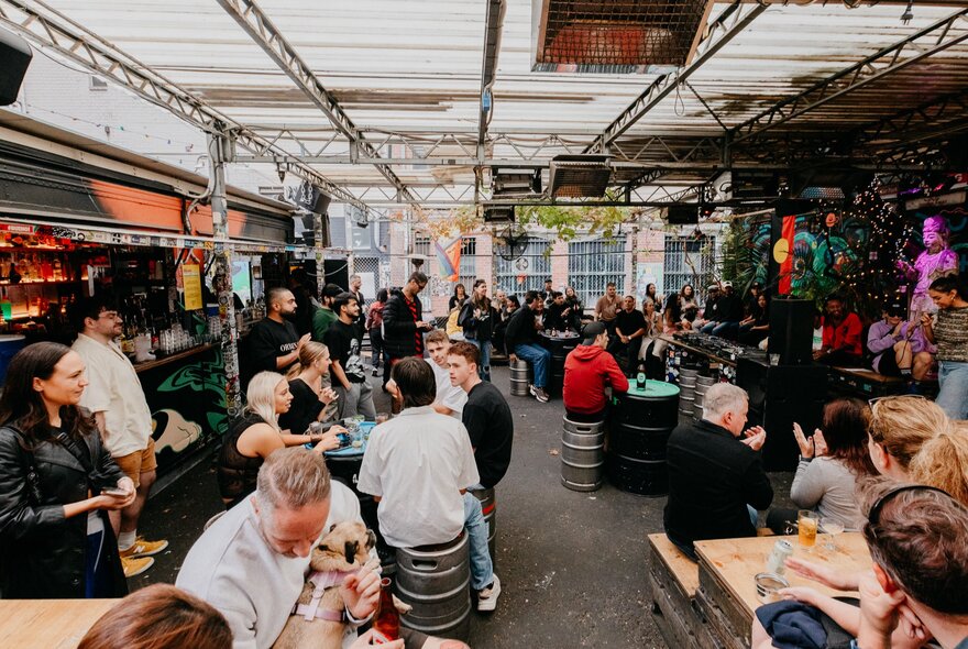 A busy outdoor bar courtyard filled with patrons sitting and standing at tables, with an industrial steel roof structure overhead, a bar counter on the left, and vibrant graffiti artwork on the right wall.
