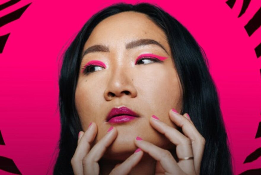 The face of comedian Charlene Kaye, with straight long black hair framing her face, pink eye shadow and pink lipstick, her fingers resting lightly on her chin area, against a hot pink background.