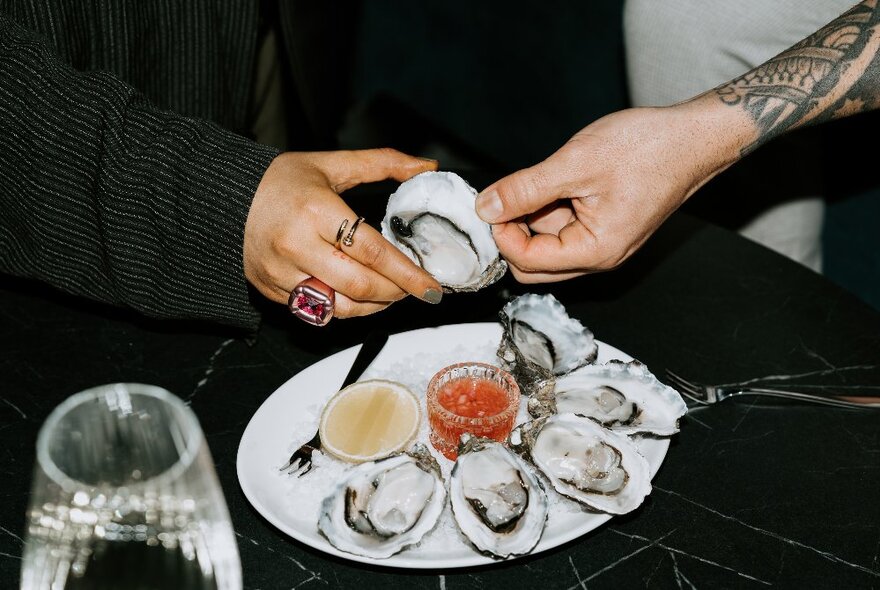 Hands reaching in towards a plate of freshly shucked oysters on a low table.