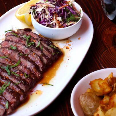 A sliced steak on a white plate, served with a colourful coleslaw, a side of smashed potatoes and a glass of red wine.