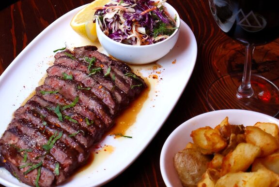 A sliced steak on a white plate, served with a colourful coleslaw, a side of smashed potatoes and a glass of red wine.