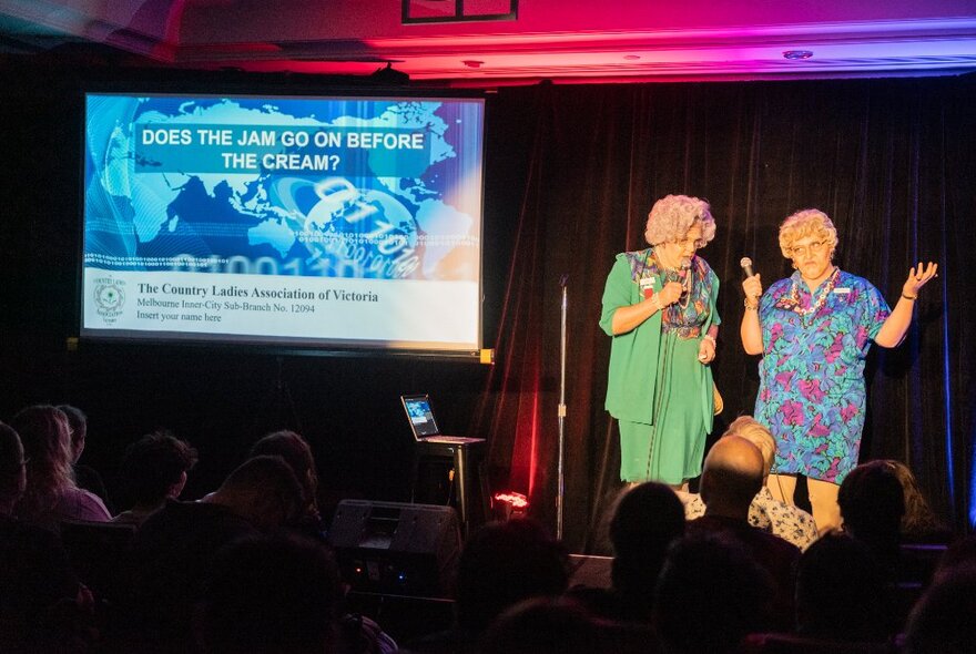 Two older female comedians on stage, with a slide presentation regarding the correct order of jam and cream on scones behind them.