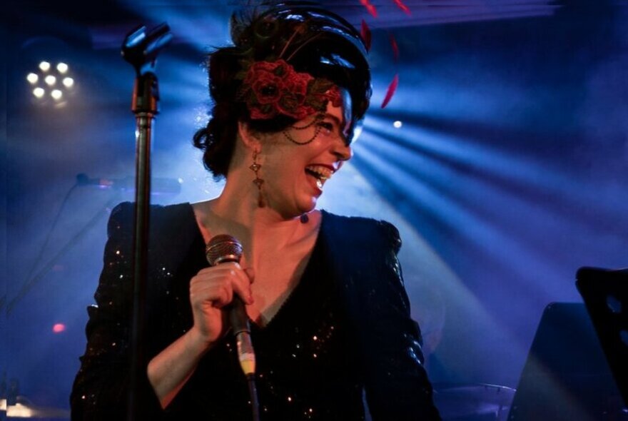 Singer wearing a cocktail hat and sequinned dress, holding the microphone and smiling on stage, with blue filtered light behind her.