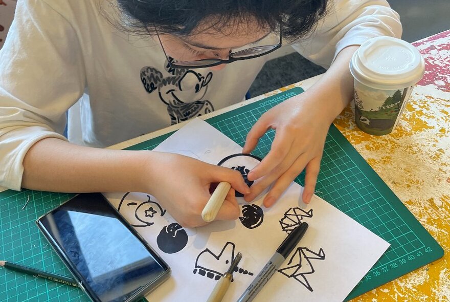 Person busy drawing with a marker pen on a white piece of paper at a work table.