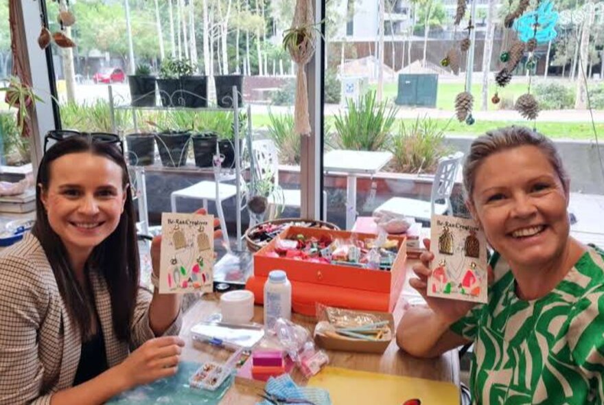 Two smiling workshop participants seated at a table with craft materials, holding up jewellery they have made.