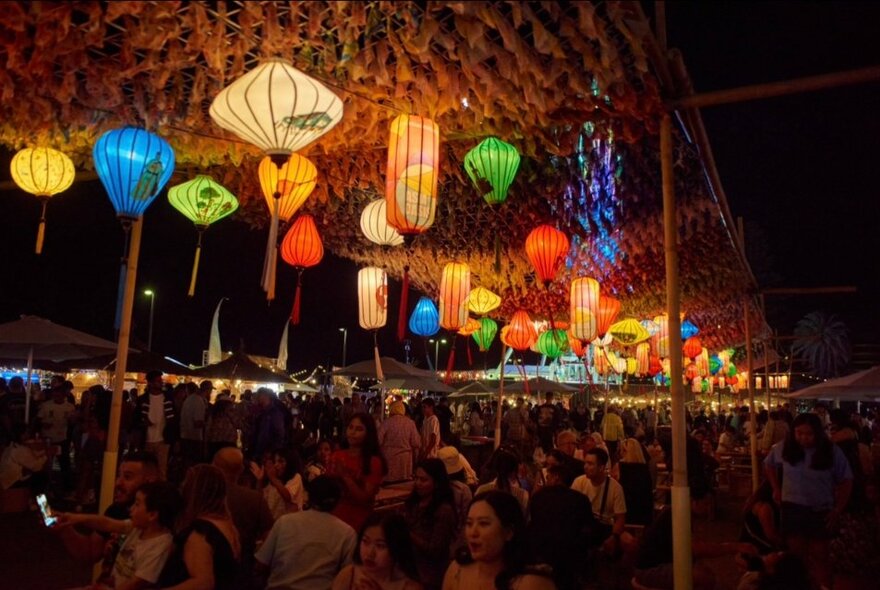 Rows of colourful Hoi An style lanterns, suspended from a permeable roof, at night, with a crowd of peopl walking around beneath them.