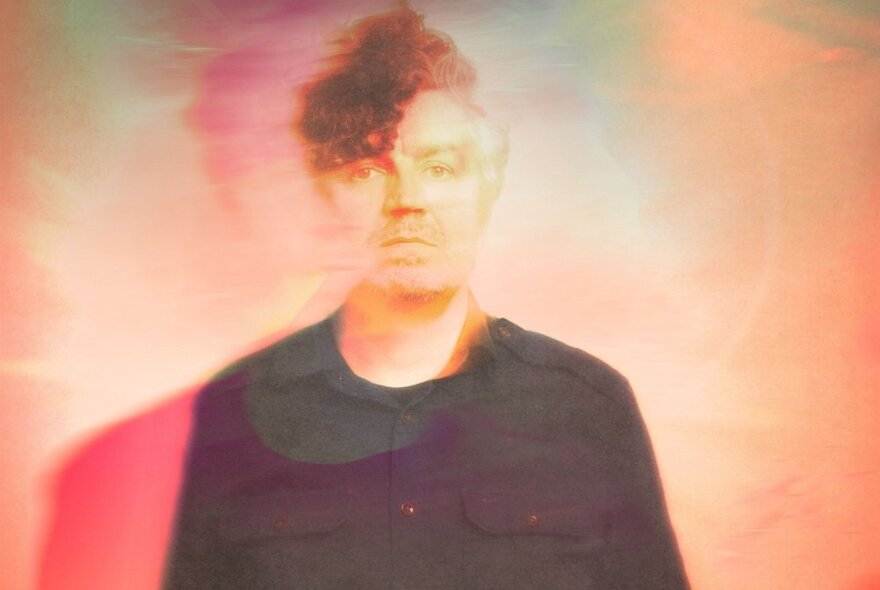 A man with dark, curly hair swept to one side, set against a hazy, multicolored background dominated by warm pink and orange tones. 