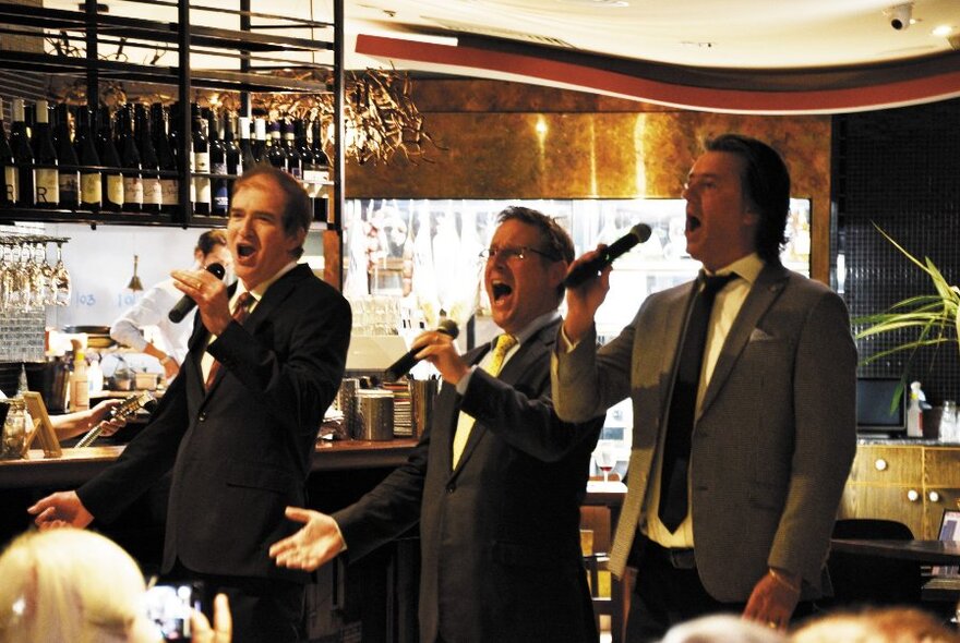 Three men wearing suits and singing into microphones, inside a restaurant to patrons seated at dining tables.