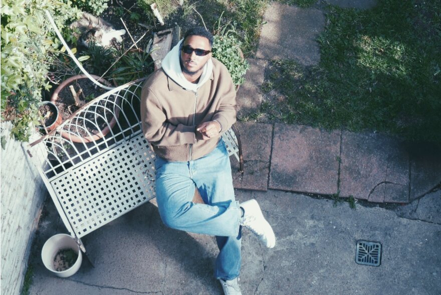 Jazz musician, DoomCannon, sitting on a metal garden seat in a paved garden, looking up at the camera.
