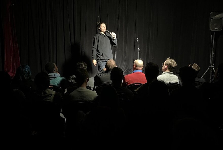 Person in dark clothing holding mic, in front of audience in darkened room.
