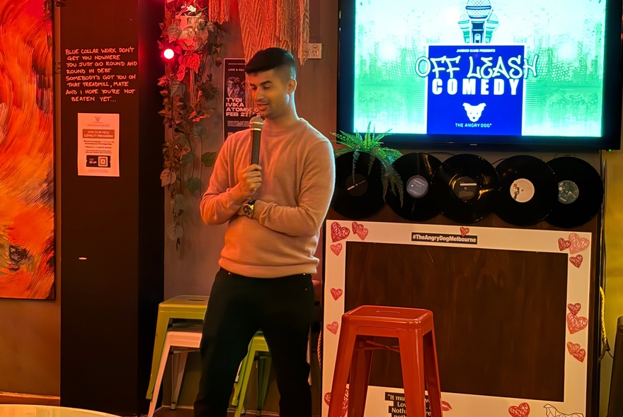 A man in a fitted brown jumper standing with a microphone in front of a presentation screen and two stools, inside a bar setting. 