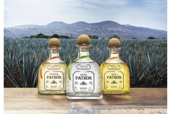 Three tequila bottles resting on a wooden table in front of an agave plantation, mountains in the distance.