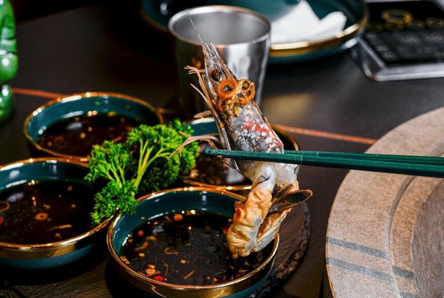 Chopsticks holding a cooked prawn over dishes of Asian sauces.