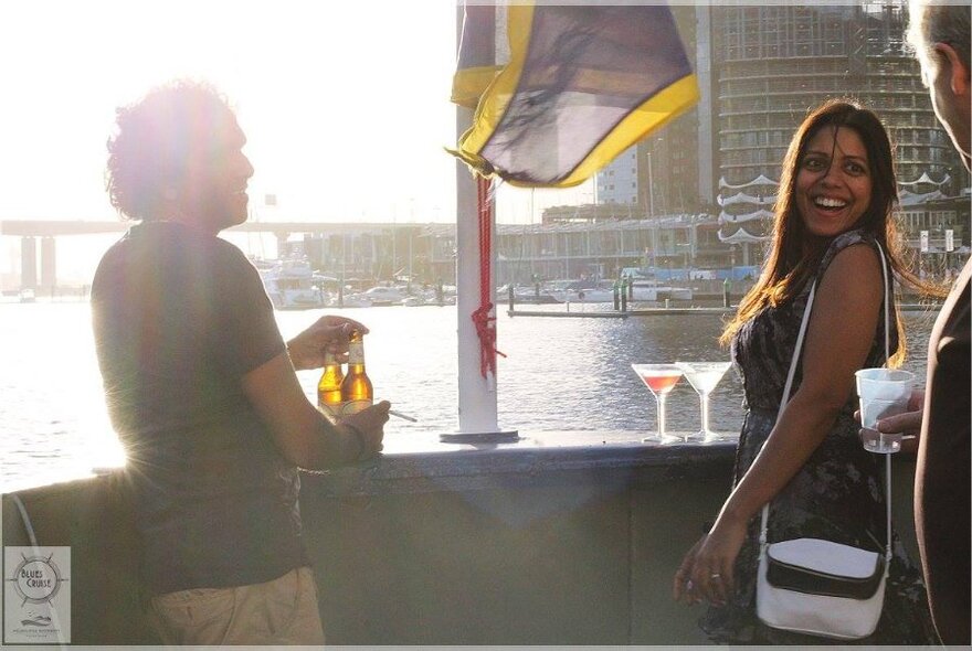 Three people at the edge of a boat, laughing,  with beverages.