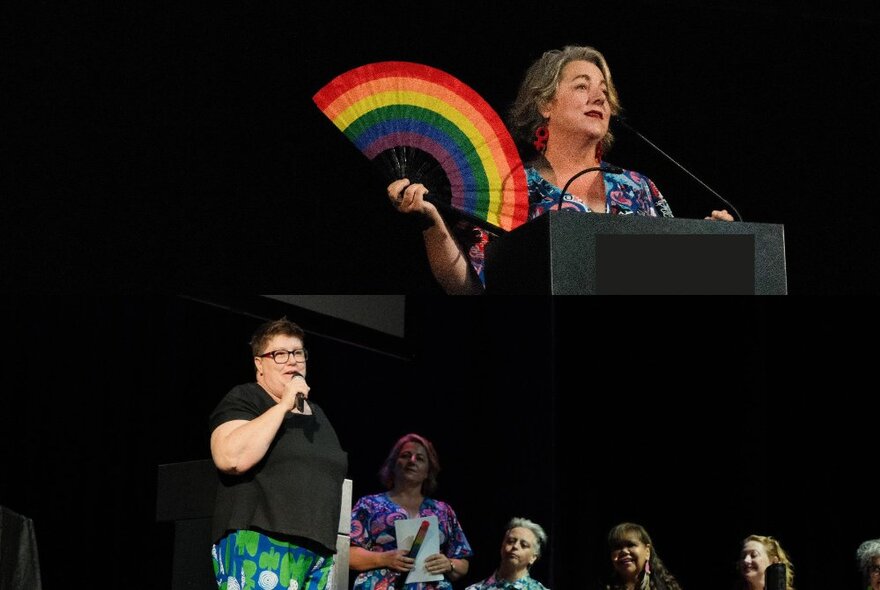 A woman at a lecturn during a debate holding a rainbow fan, people visible in the background. 