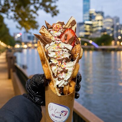 A gloved hand holding up a loaded creamy Belgian sweet waffle in a cone, against a backdrop of the Yarra River and Melbourne city at evening twilight.