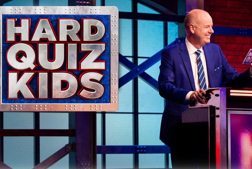 Comedian Tom Gleeson standing behind a lectern, smiling, on the set of the TV show Hard Quiz Kids, with a large sign behind him with those words on it.