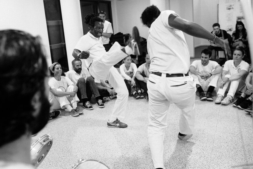 A black and white image of people demonstrating the Afro-Brazilian martial art of Capoeira in a small room with people sitting on the floor looking on. 