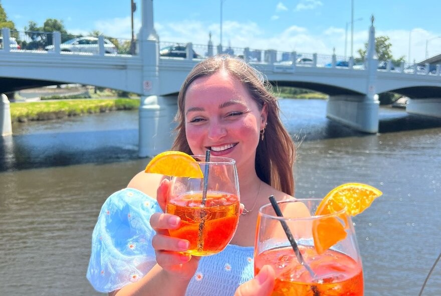 A woman smiling on the Yarra River with a bridge in the background while holding an Aperol spritz, another Aperol spritz in the foreground. 