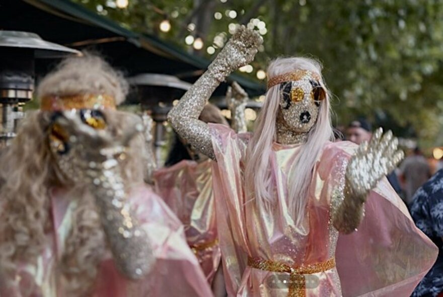 Figures wearing silver capes and bodysuits, in an outdoor leafy setting.