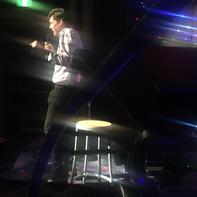 A blurred photo of a person on stage with a stool, doing a stand-up comedy routine.