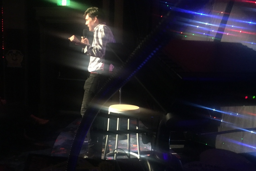 A blurred photo of a person on stage with a stool, doing a stand-up comedy routine.