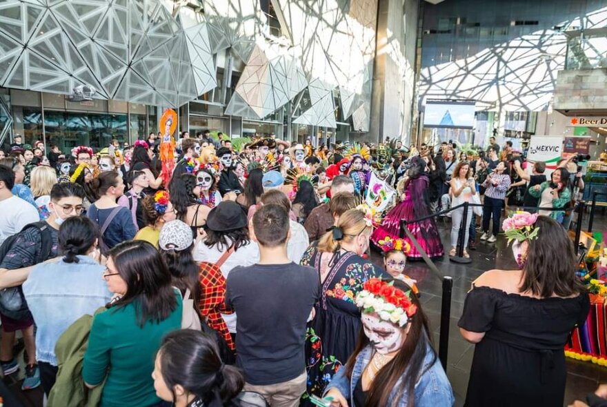 Day of the Dead celebrations taking place at the Atrium in Fed Square, with a large crowd of people milling around.