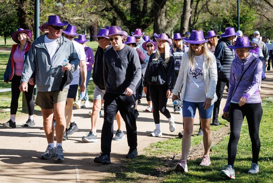 Charity walk teams wearing purple cowboy hats and running gear following a gravel path in a parkland setting.