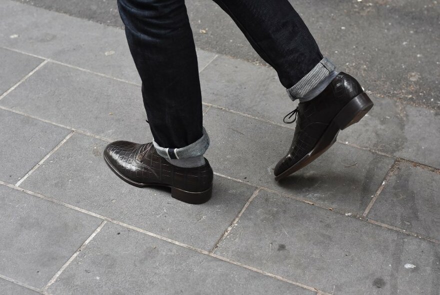 Detail of man's legs wearing jeans and lace-up leather shoes on a street pavement.