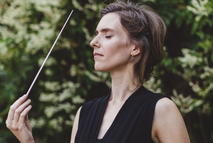 Conductor Chloé van Soeterstède standing side-on with her eyes closed and baton raised in a garden setting. 