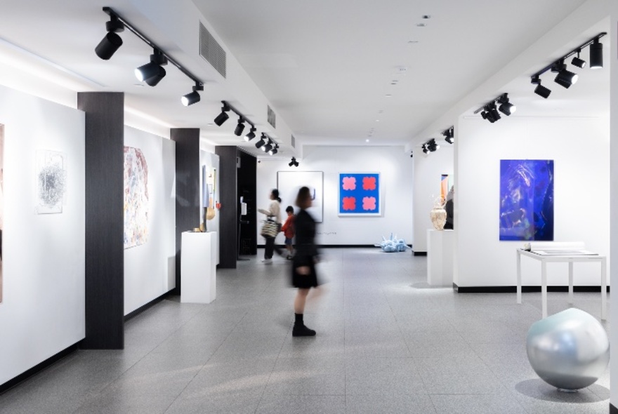 White-walled gallery space hung with large artworks and lit by rows of overhead spotlights, blurred person in the centre.