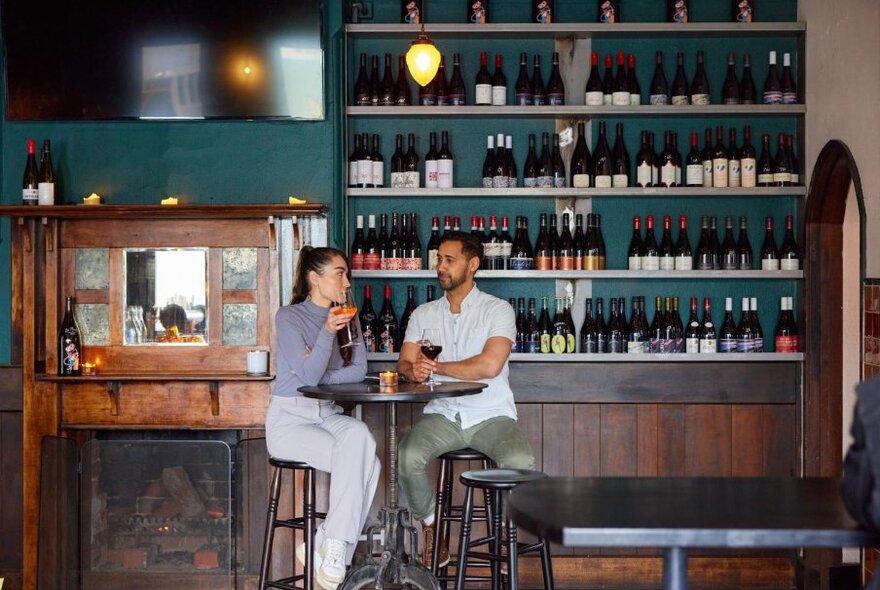 Two people at a tall table and stools in front of a wall with wine bottles