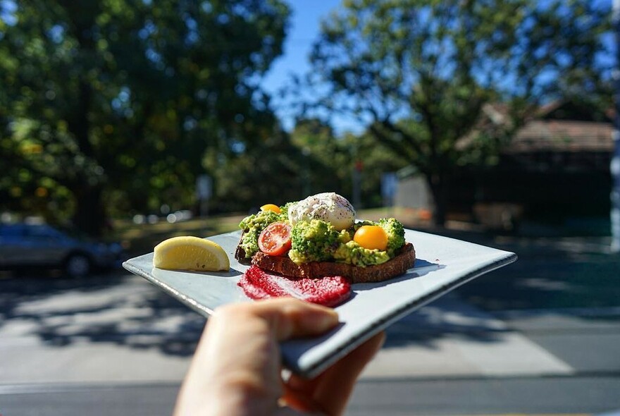 A hand holding a square white plate with avocado and poached egg on toast.