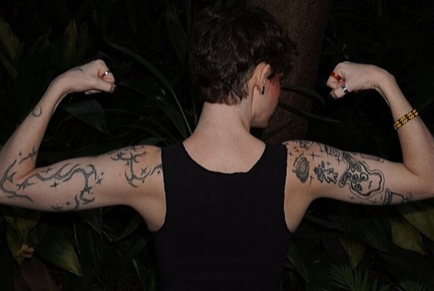 Boy with tattoos and short black top, flexing his muscles, seen from behind.