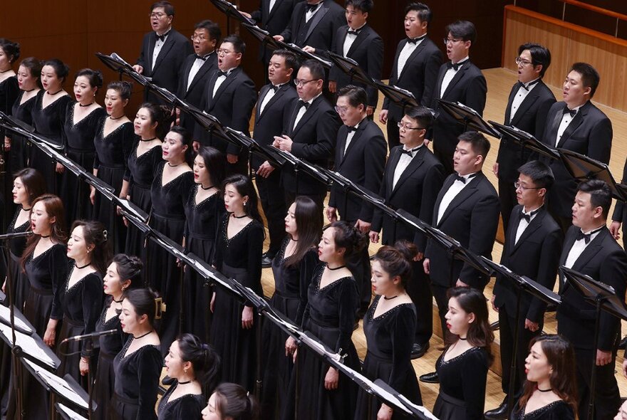 Members of the China National Symphony Orchestra Chorus singing on a stage, the women wearing black dresses, the men in tuxedos, all of them standing in front of individual music stands.